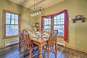 A dining room with a wooden table set for six, flanked by two windows with blinds, a chandelier, and a clock on the wall.