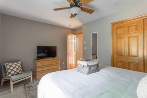 Quarto com cama, televisão, penteadeira, um banco, mala, ventoinha de teto e porta.