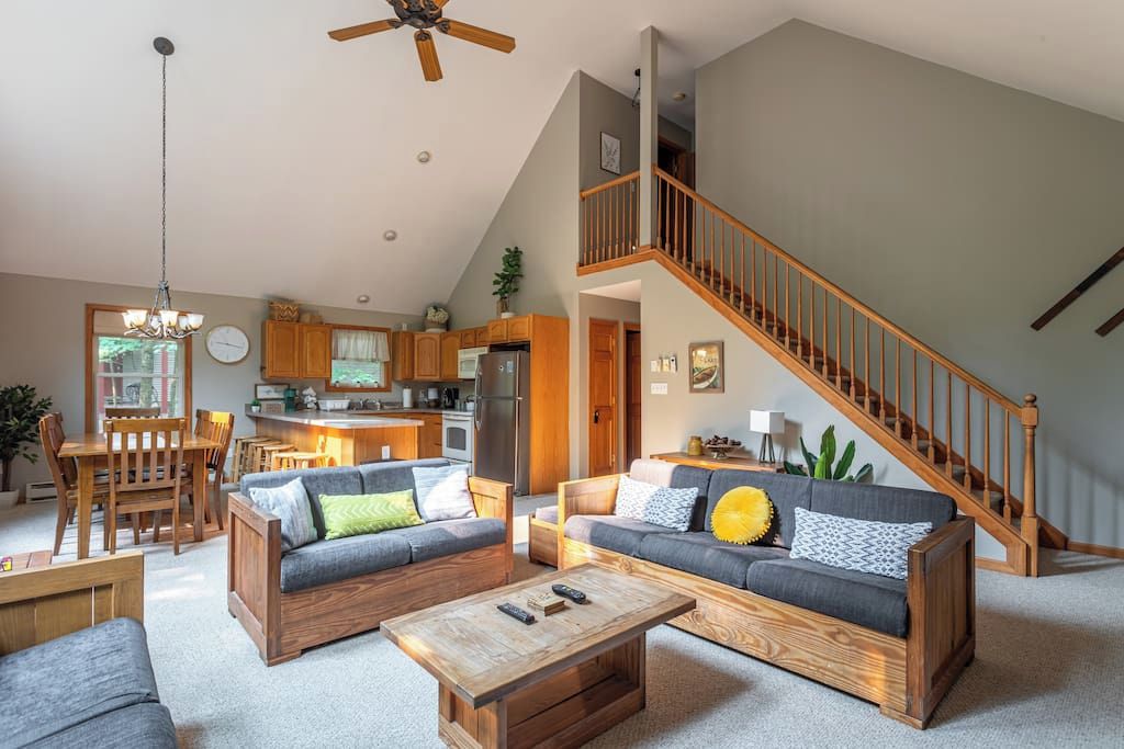 A spacious living area with a high ceiling, a wooden staircase, and a kitchen in the background.