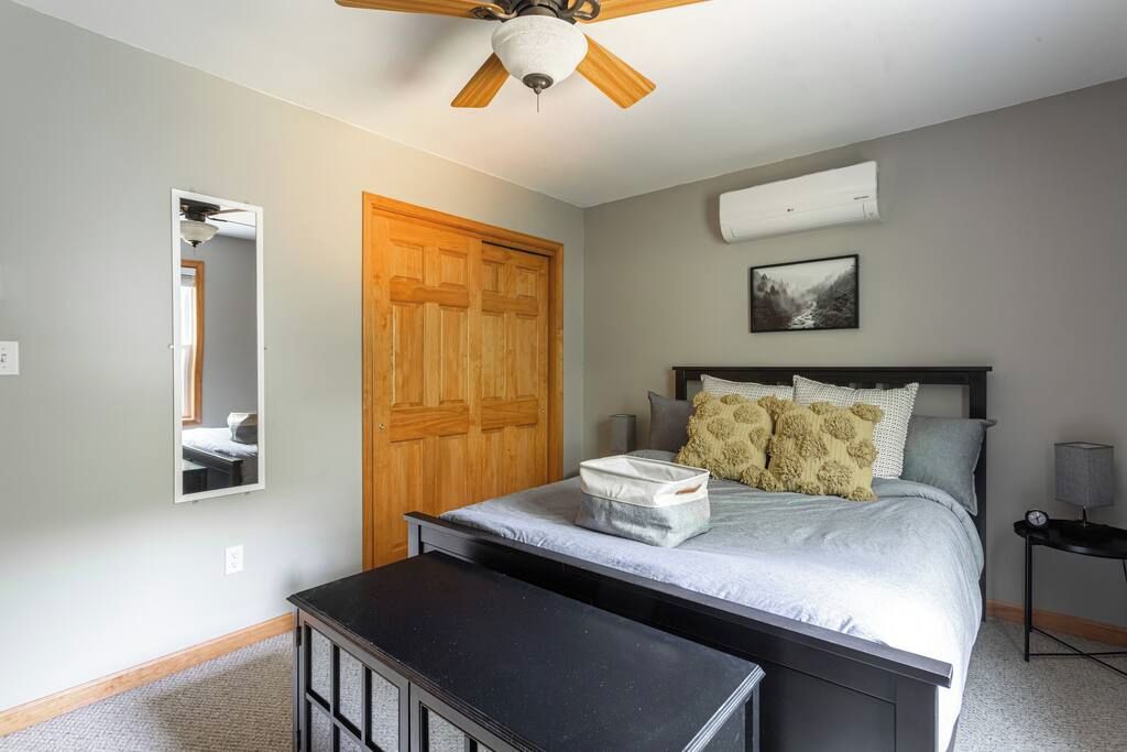 A bedroom with a bed, a wooden wardrobe, a ceiling fan, a wall-mounted air conditioner, and a bedside table.