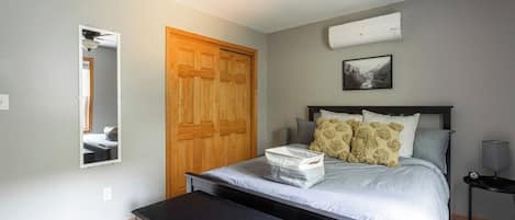 A bedroom with a bed, a wooden wardrobe, a ceiling fan, a wall-mounted air conditioner, and a bedside table.
