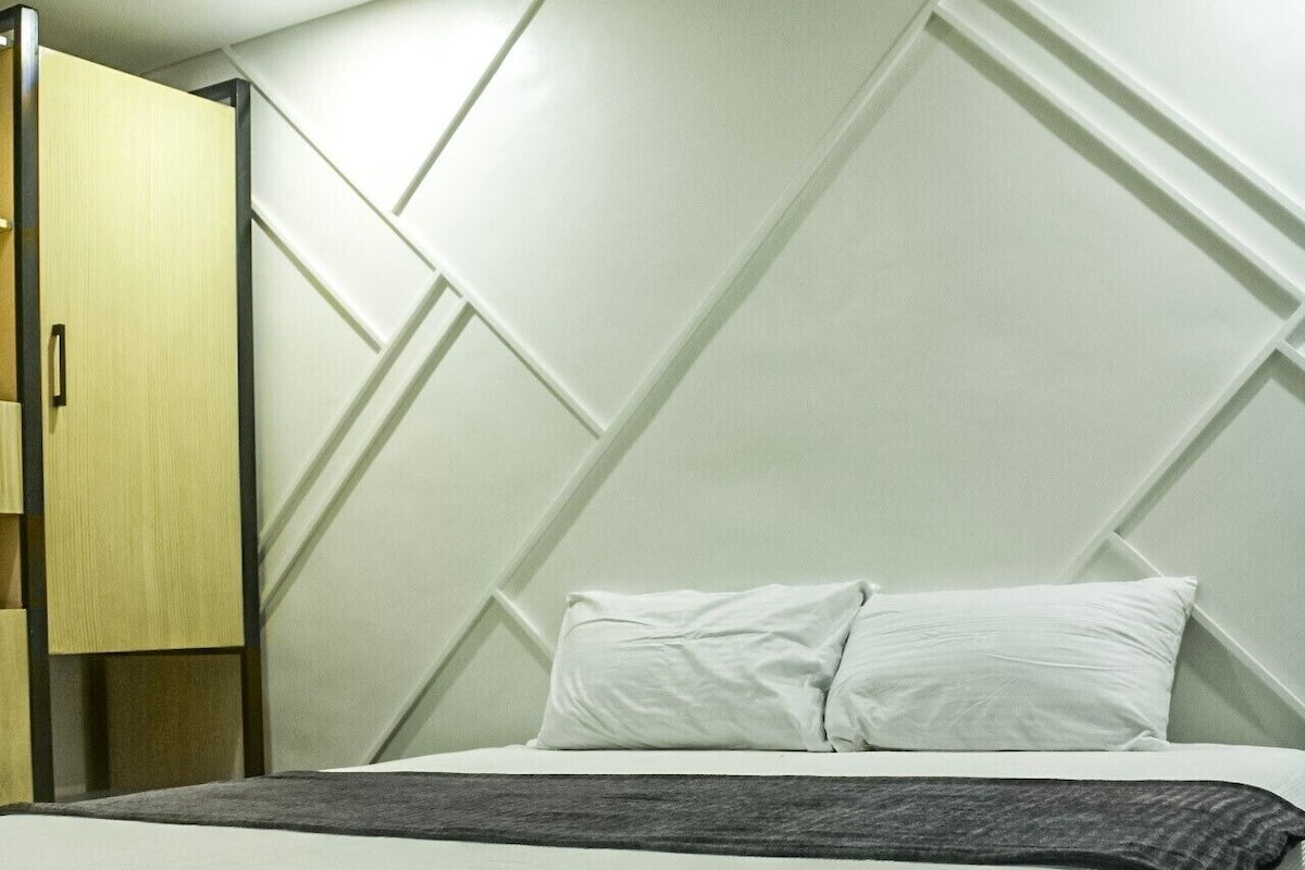 A neatly made bed with a grey blanket, a wooden wardrobe, and a geometric wall design.