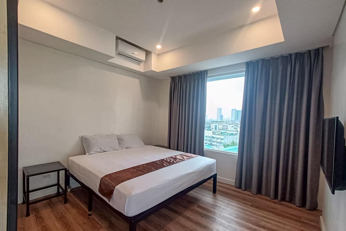 A modern hotel room with a large bed, a small bedside table, a built-in air conditioning unit, and a city view through the window.