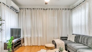 A modern living room with a grey sofa, a round coffee table, a TV on a wooden stand, and sheer curtains.