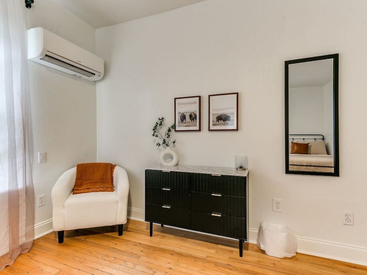 A room with a white armchair, a black dresser, and a mirror on the wall.