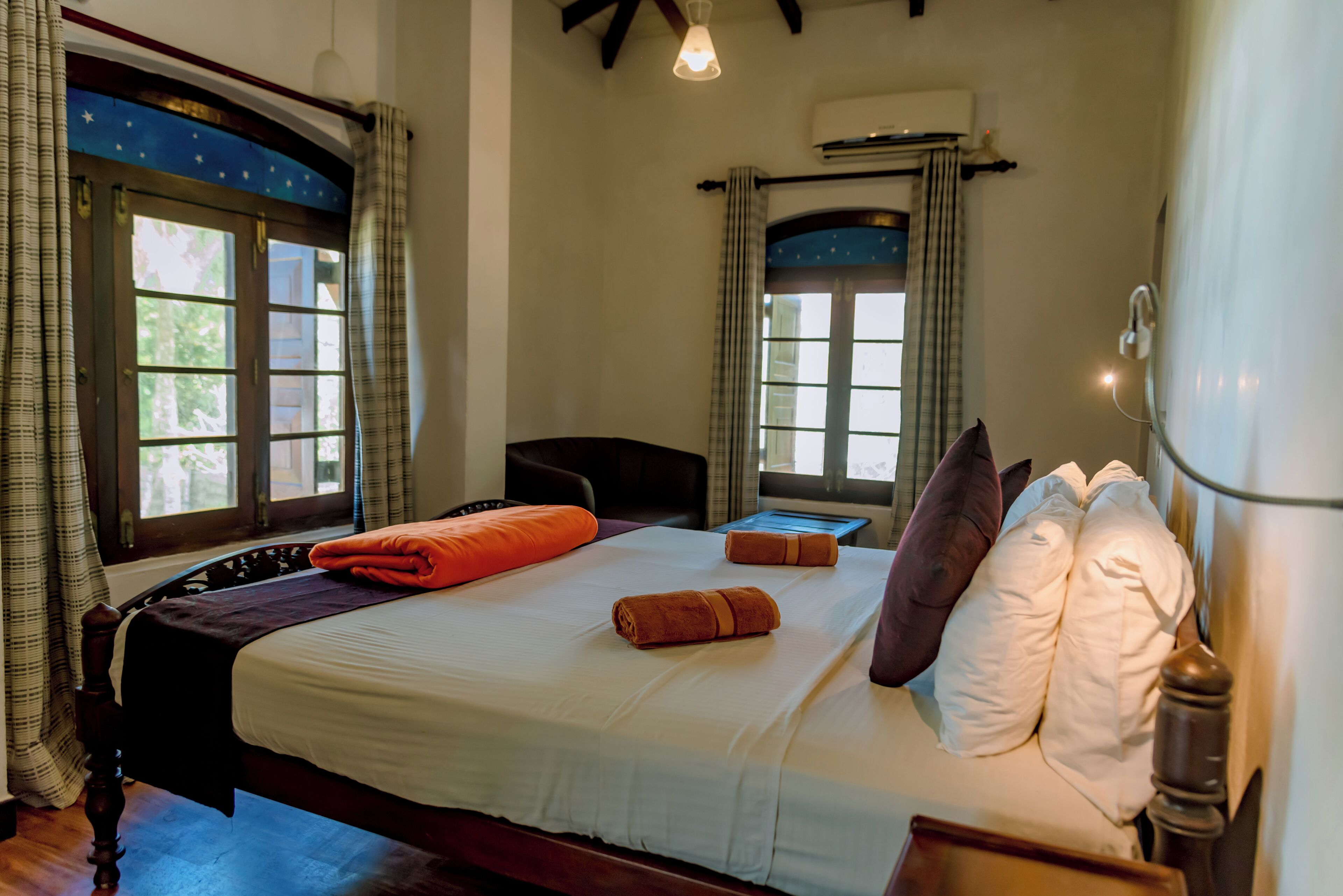 A neatly made bed with white linens and orange towels, a wooden headboard, and a window with curtains.
