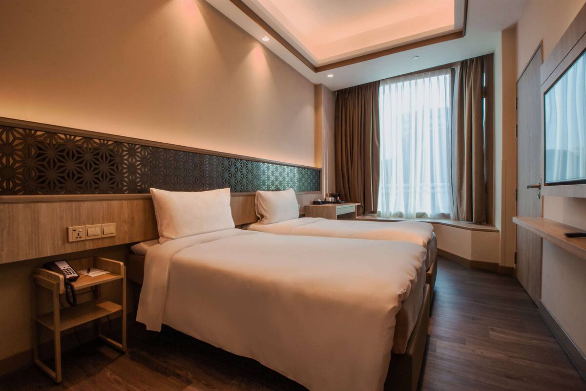 Ein Hotelzimmer mit zwei Betten, einem Nachttisch mit Telefon, einem Fenster mit Vorhängen und einer Wand mit einer Zierleiste.
