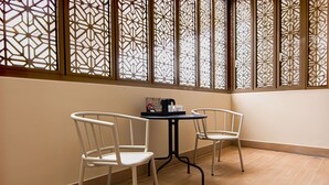 Une pièce avec deux chaises blanches et une petite table ronde, une fenêtre décorative ornée de motifs complexes, et un sol carrelé de couleur claire.