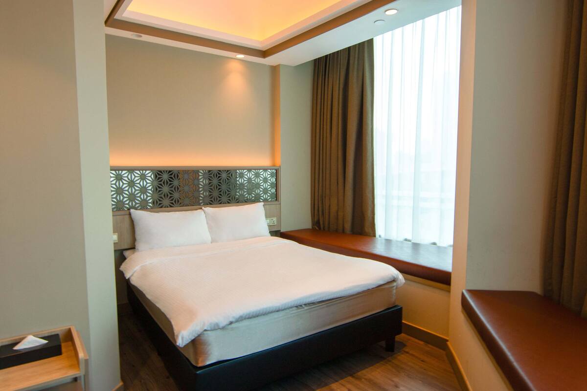 Ein Hotelzimmer mit einem großen Bett, einem Nachttisch, einem Fenster mit Vorhängen und einem dekorativen Kopfteil.