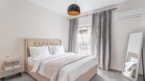A bedroom with a large bed, a bedside table with drawers, a hanging light fixture, a window with curtains, and an air conditioning unit.