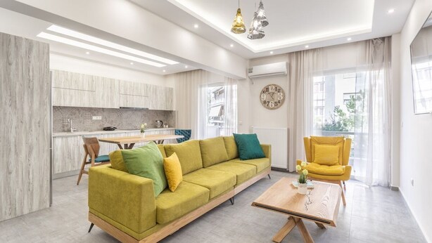 A modern living room with a sofa, coffee table, and yellow armchair. A kitchen is visible in the background with a dining area.