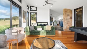 Ein modernes Wohnzimmer mit einem grünen Sofa, einem weißen Sofa und einem Billardtisch. Große Fenster bieten einen Blick ins Freie.