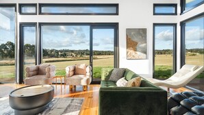 Ein modernes Wohnzimmer mit großen Fenstern, einem grünen Sofa, einem beigefarbenen Sessel und einem Liegestuhl.