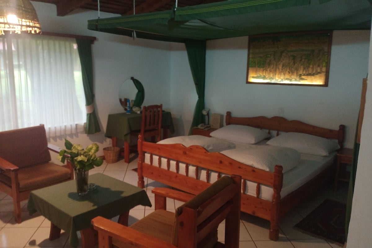 A room with a bed, a wooden chair, a small table, and a hanging lamp.