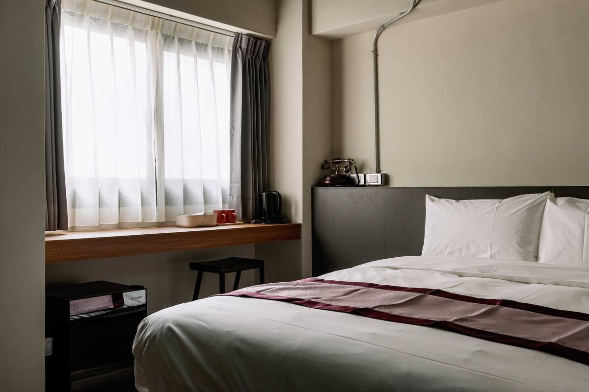 A hotel room with a bed, a desk with a chair, a window with curtains, and a nightstand with a lamp.