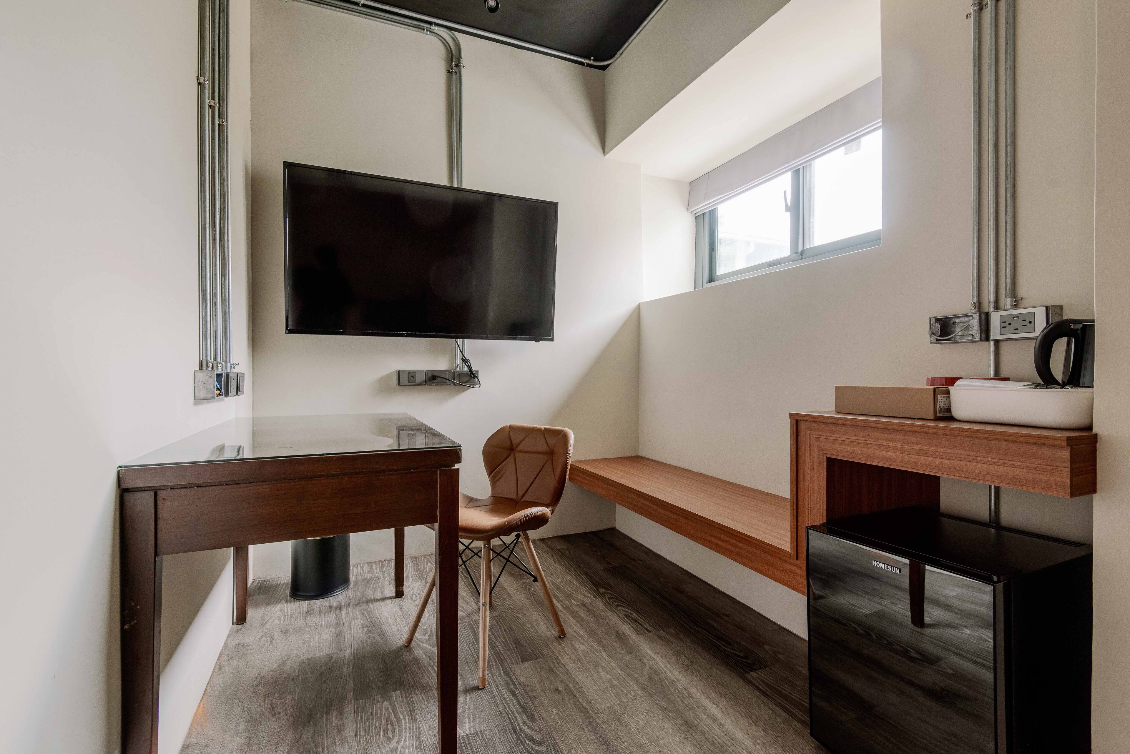 Une chambre moderne et compacte, dotée d’un téléviseur à écran plat fixé au mur, d’un bureau en bois, d’une chaise rembourrée, d’une banquette intégrée et d’un réfrigérateur noir.