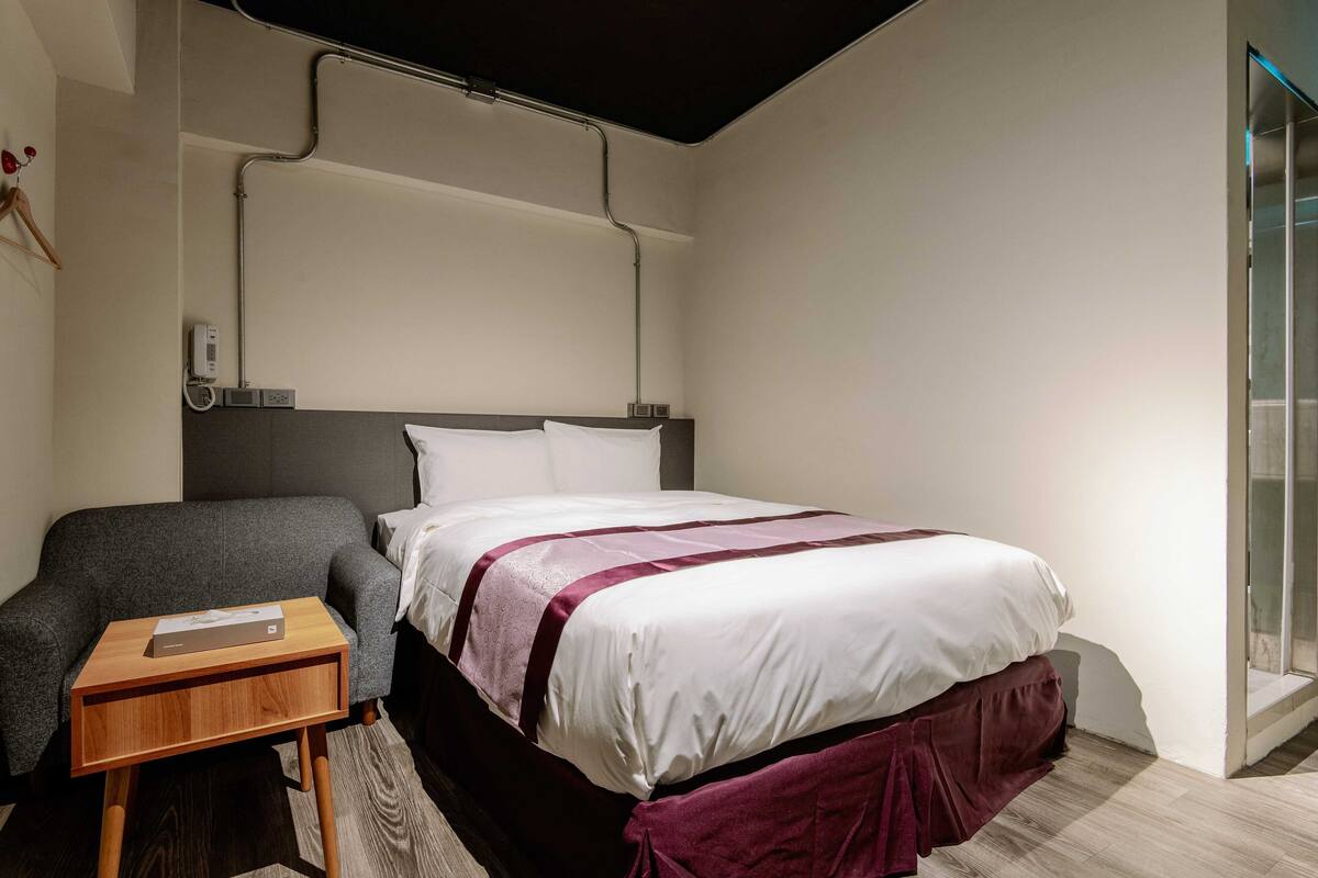 A modern hotel room with a bed, a bedside table, a chair, and a glass shower enclosure.