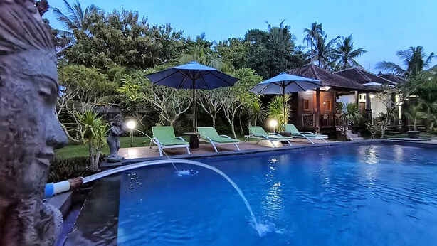 Una piscina con sillones de descanso y sombrillas, rodeada de vegetación tropical y un edificio al fondo.