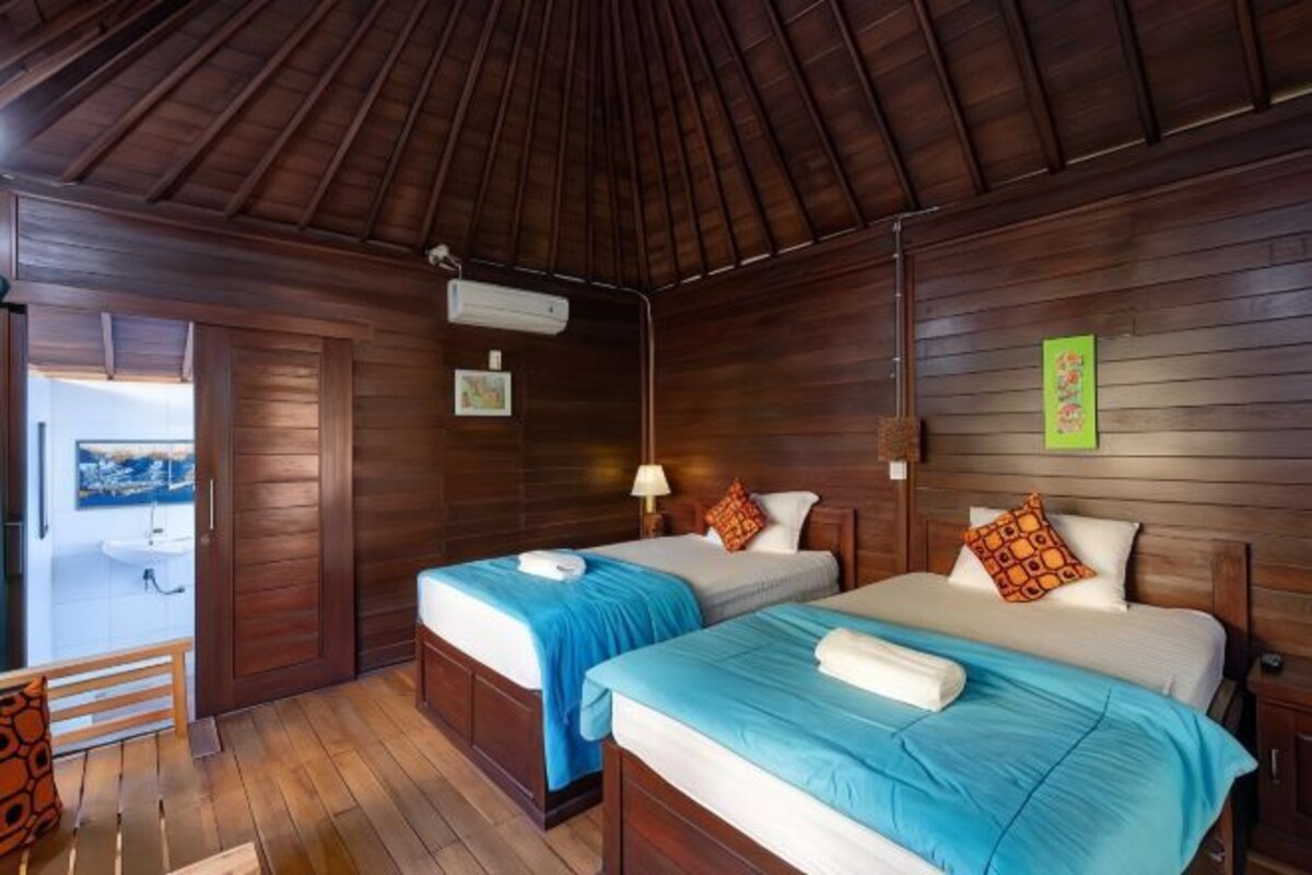 Quarto com duas camas, paredes de madeira, ventilador de teto e uma pequena mesa.