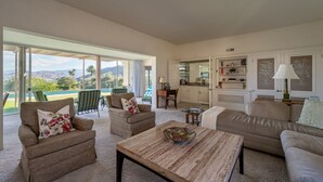 A spacious living room with a large window overlooking a pool and mountains, furnished with a sofa, chairs, and a coffee table.