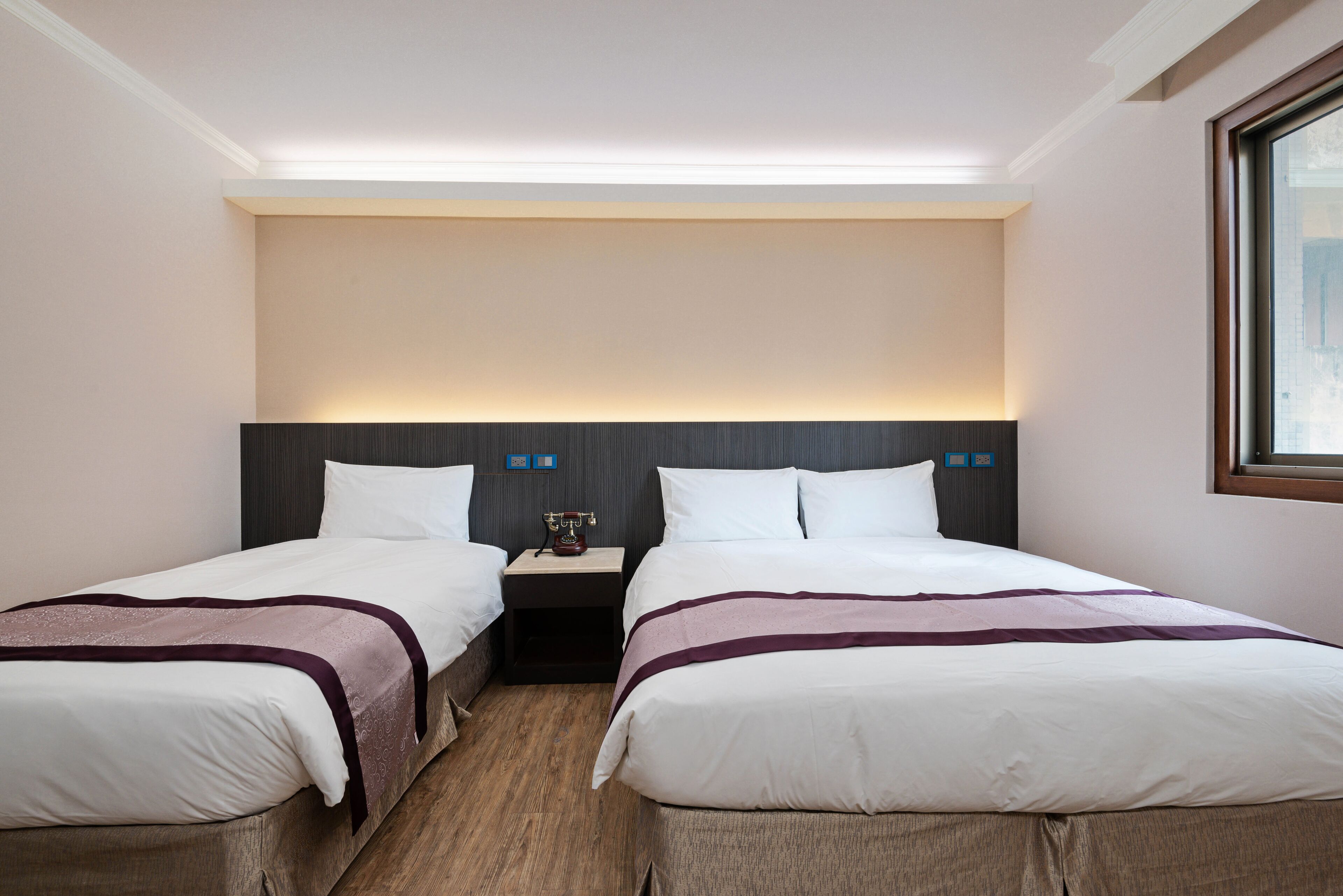 A hotel room with two beds, a nightstand, and a wall-mounted lamp.