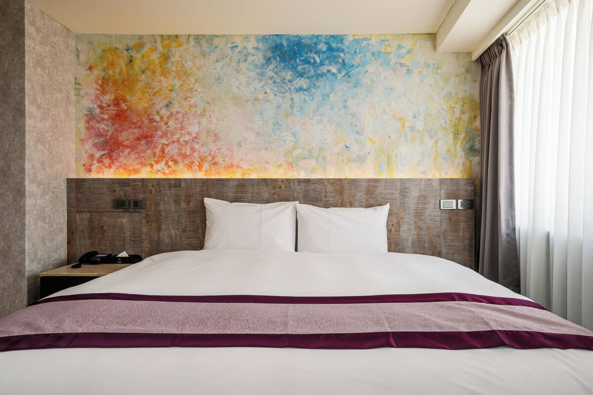 A hotel room with a large bed, a wooden headboard, a nightstand with a phone, and a colorful abstract wall.