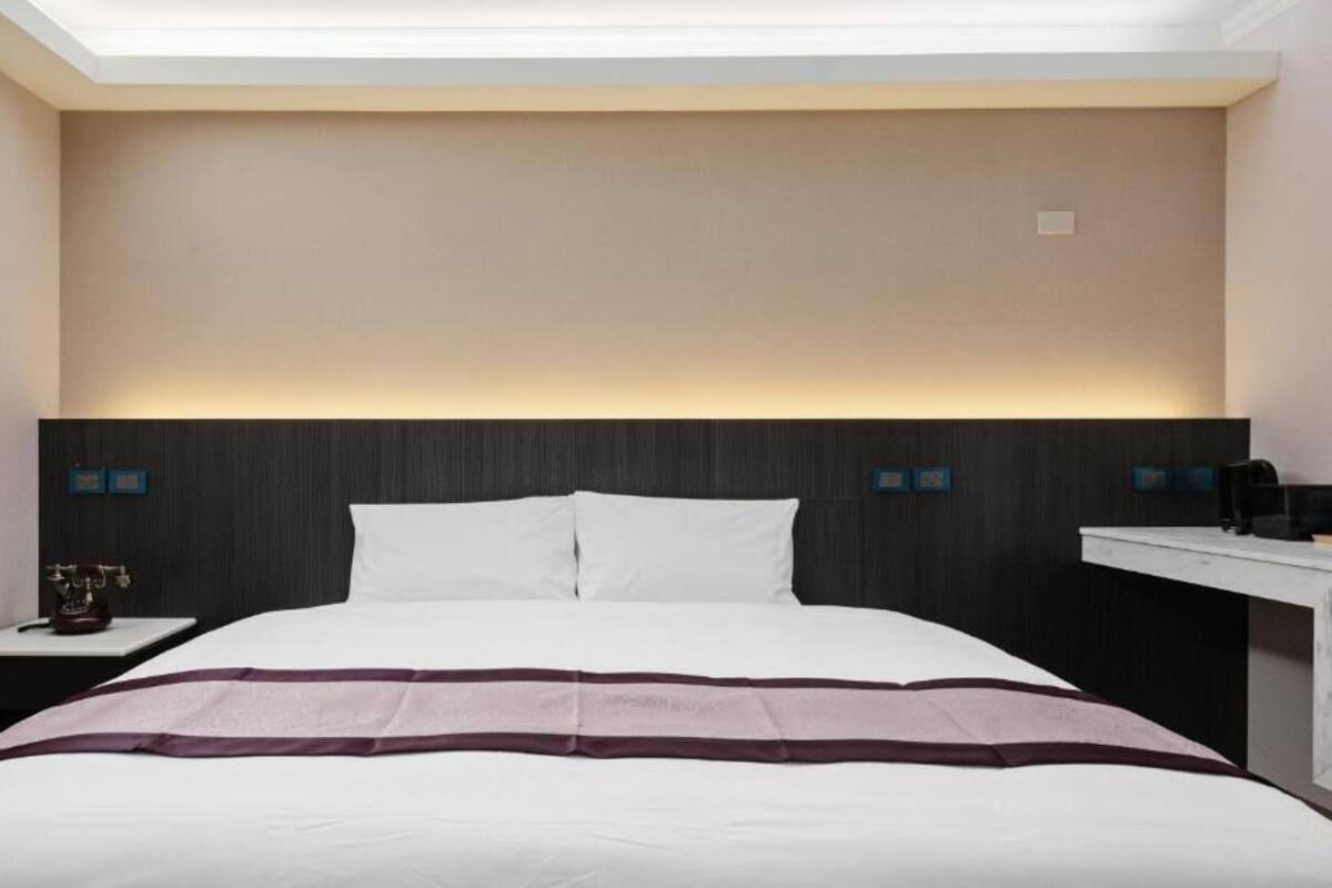 A modern hotel room with a large bed, a bedside table, a nightstand, and a desk with a chair.