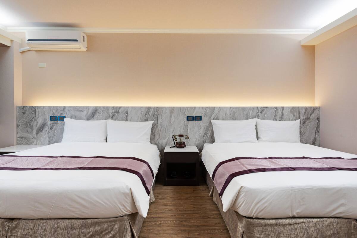 A hotel room with two beds, marble headboard, bedside tables, and an air conditioning unit.