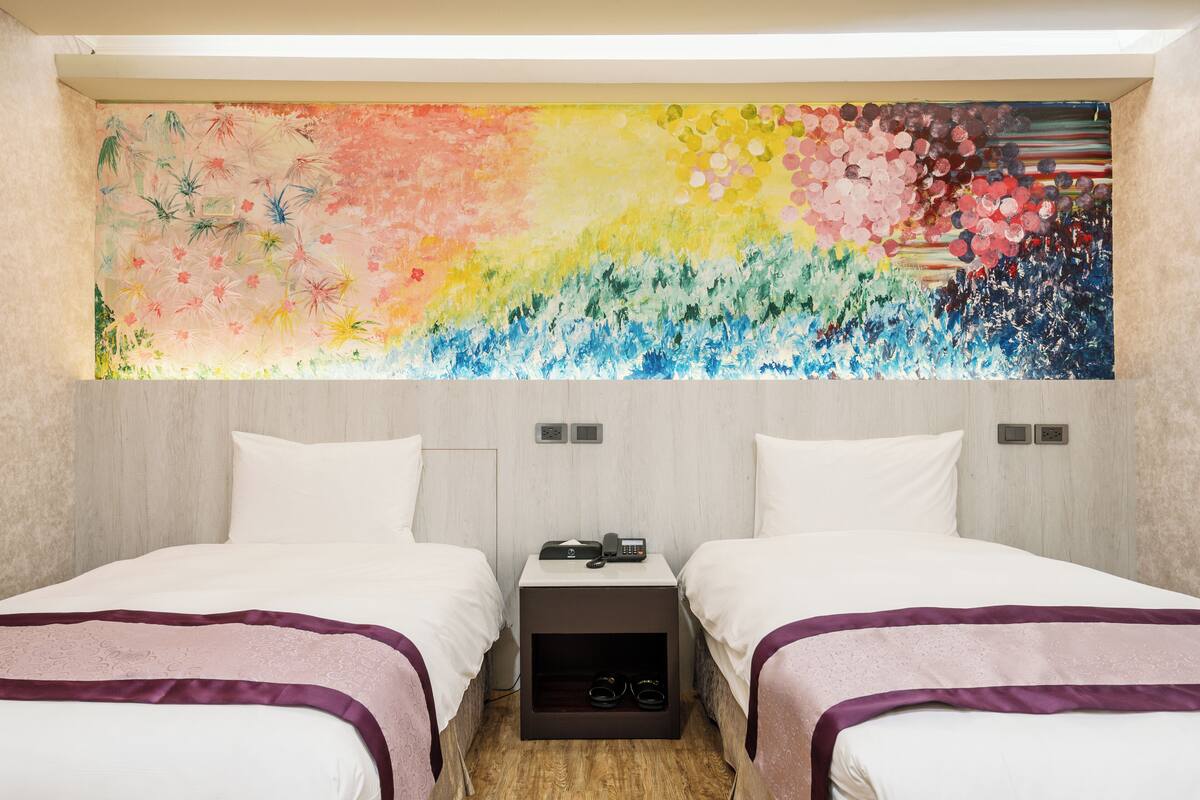 A hotel room with two beds, a nightstand with a phone, and a colorful abstract painting above the beds.