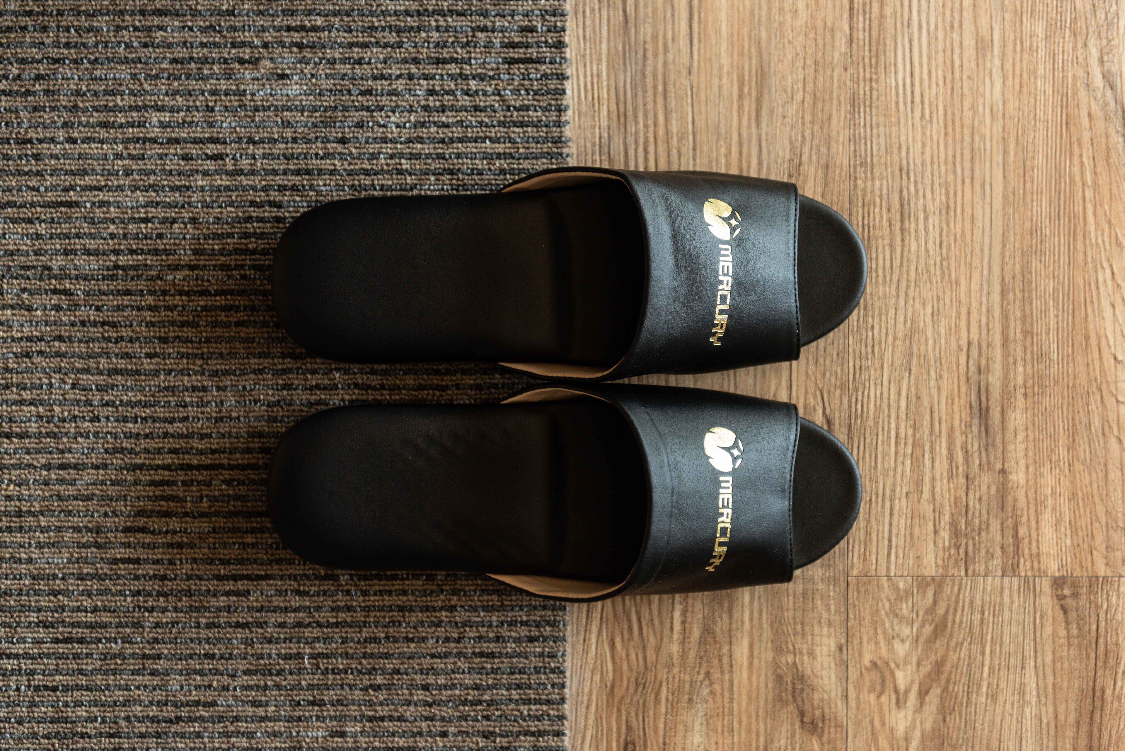 A pair of black slippers with a logo on the side, placed on a carpeted floor next to a wooden floor.