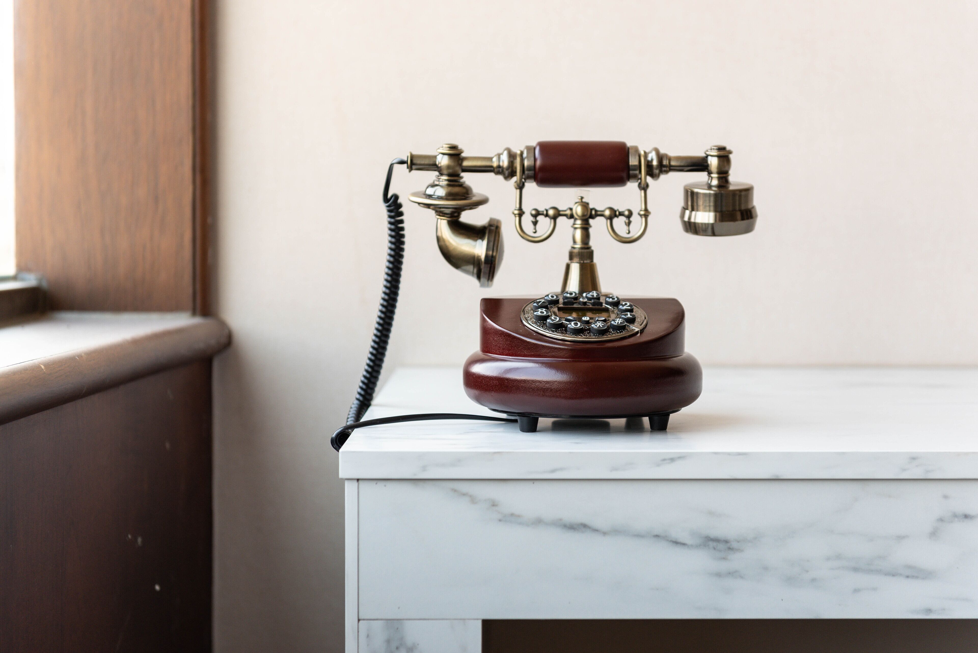 Un vieux téléphone à cadran rotatif, posé sur une table surmontée d’un plateau en marbre, dans une pièce dotée d’une porte en bois et d’un mur sobre.