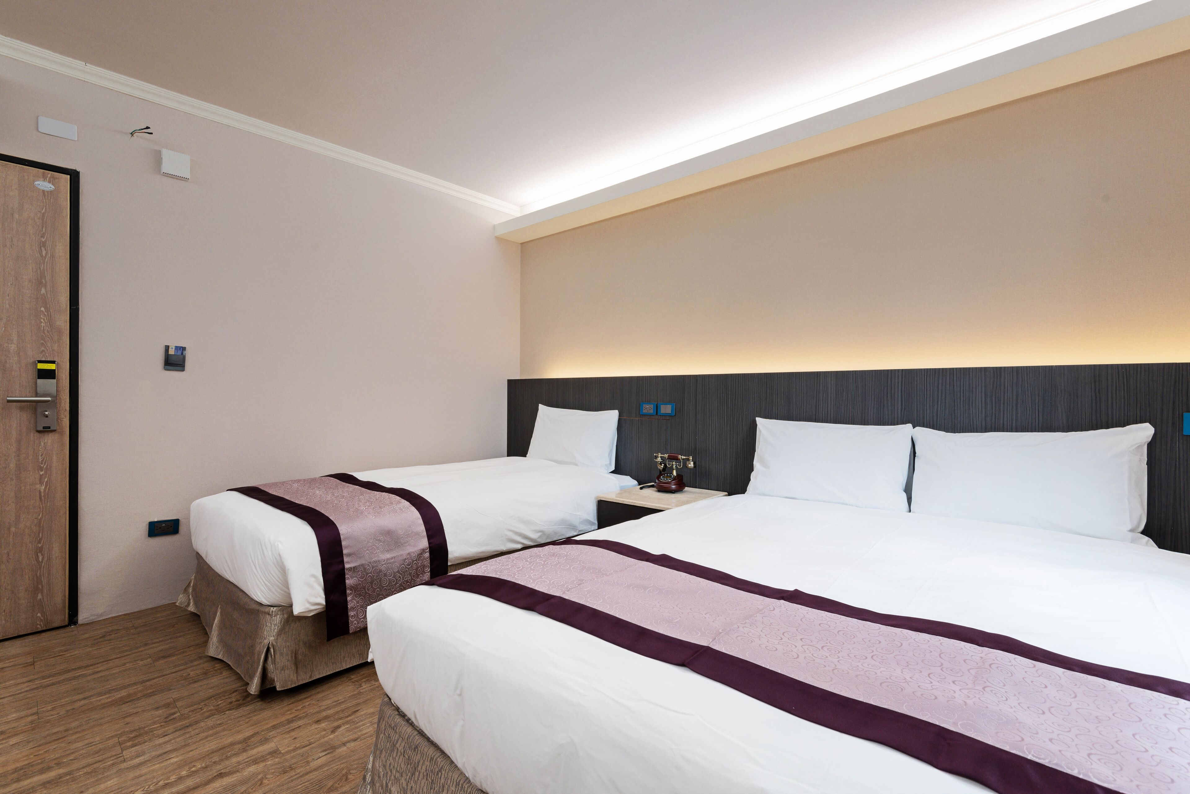 A hotel room with two beds, a wooden door, and a nightstand with a lamp.