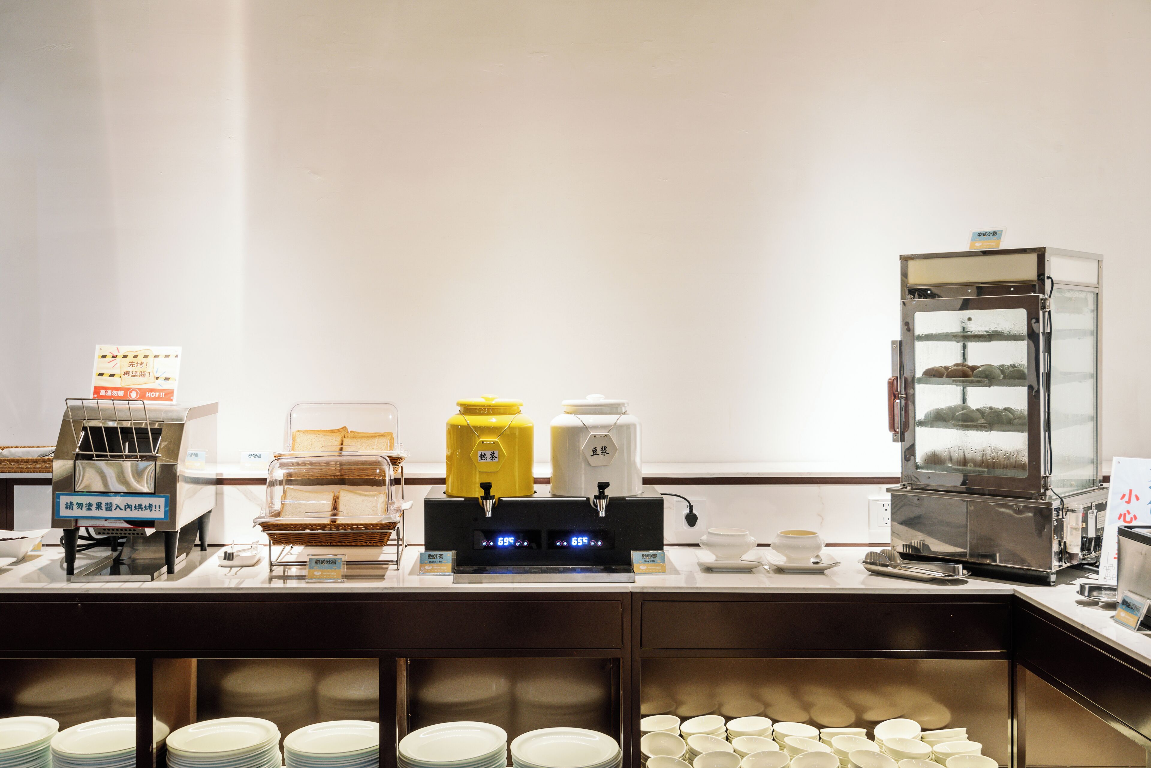 A buffet setup with a coffee machine, a food display case, and stacked plates and bowls.