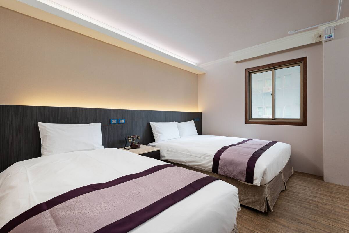 A hotel room with two beds, a nightstand, a window, and a ceiling light.
