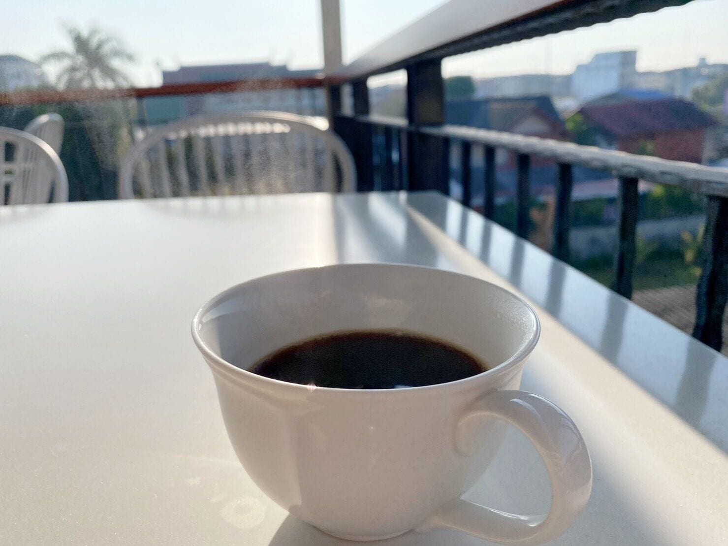 Una taza de café blanco sobre una mesa de balcón con vistas a edificios y una palmera.