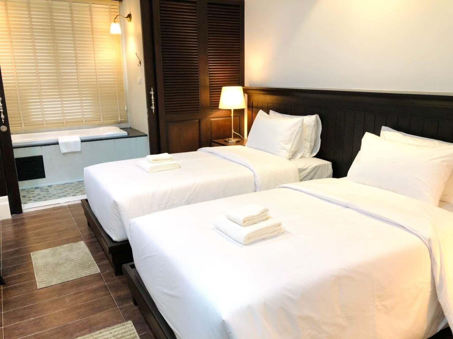 A hotel room with two beds, a window with blinds, a nightstand with a lamp, and a wooden headboard.