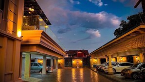 Entrada de hotel con un paseo techado, coches estacionados y cielo despejado.
