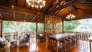 A spacious wooden deck with tables and chairs, a large chandelier, and a view of greenery.