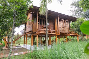 Une maison en bois dotée d’un balcon, entourée d’herbes hautes et d’arbres.