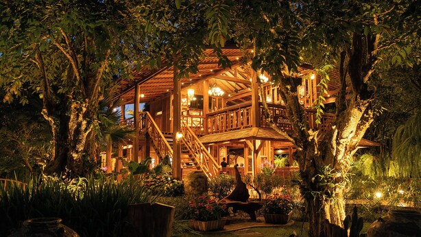 A large, rustic wooden building with multiple levels, surrounded by lush greenery and potted plants.