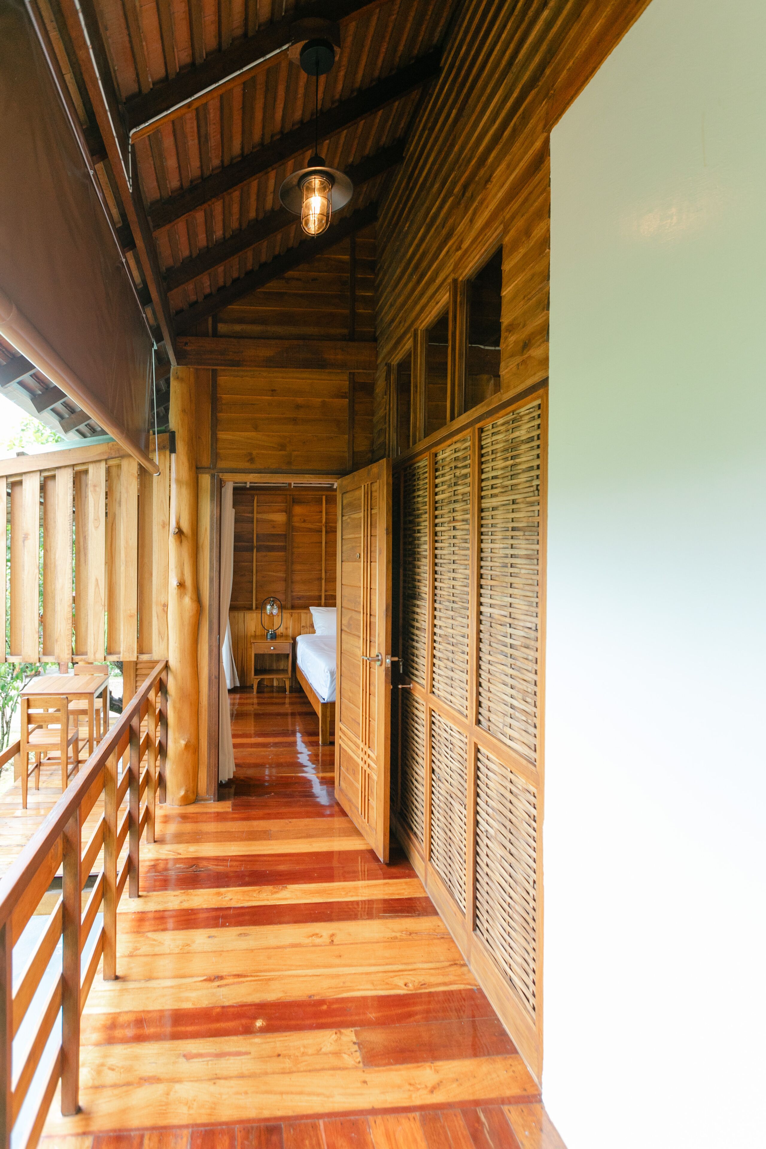A wooden corridor with a wooden floor, a bed with a white mattress, a nightstand with a mirror, and a hanging light fixture.