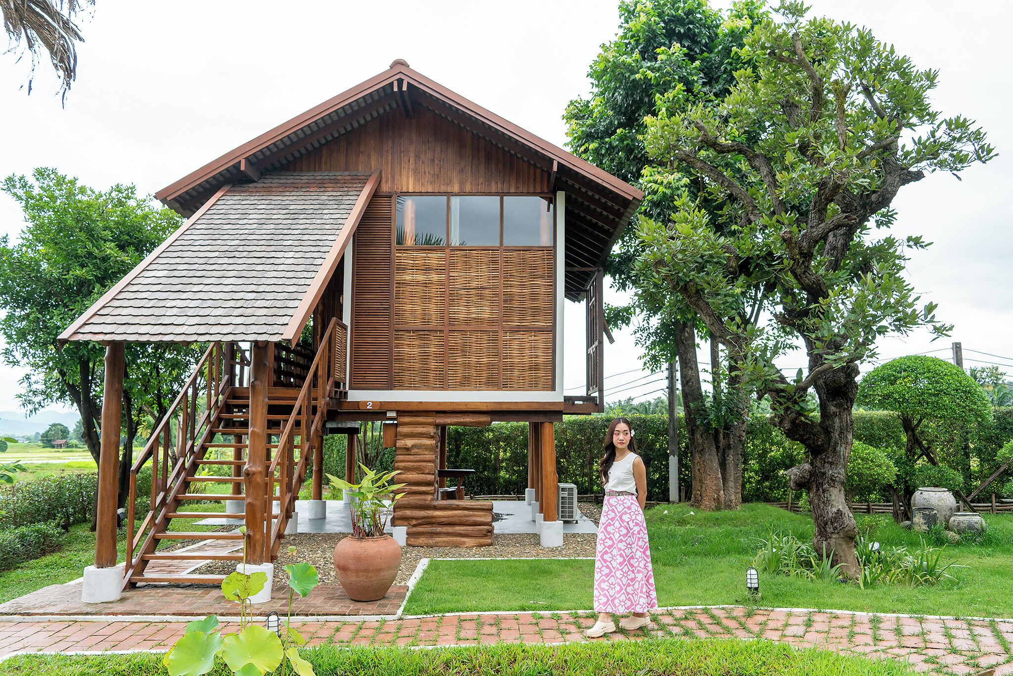 A wooden house with a staircase leading to a balcony, surrounded by lush greenery and trees.