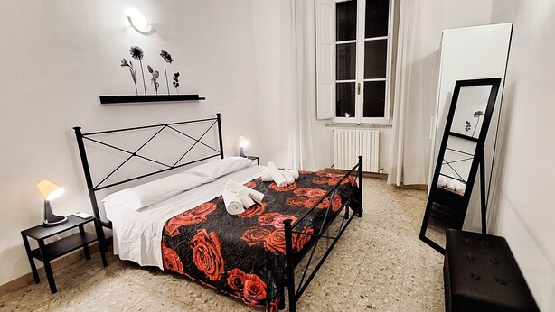 Un dormitorio con una cama, una mesita de noche con lámpara, una ventana, un radiador y un espejo.