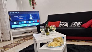 Una sala de estar con un sofá negro, cojines rojos y negros, una mesa de centro blanca con copas de vino y una botella, y un televisor mostrando un programa.