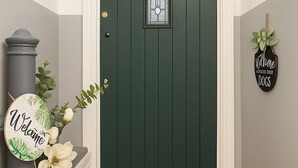 Una puerta verde oscuro con un tragaluz esmerilado, un letrero de bienvenida, una corona decorativa y un cartel que da la bienvenida a los perros.