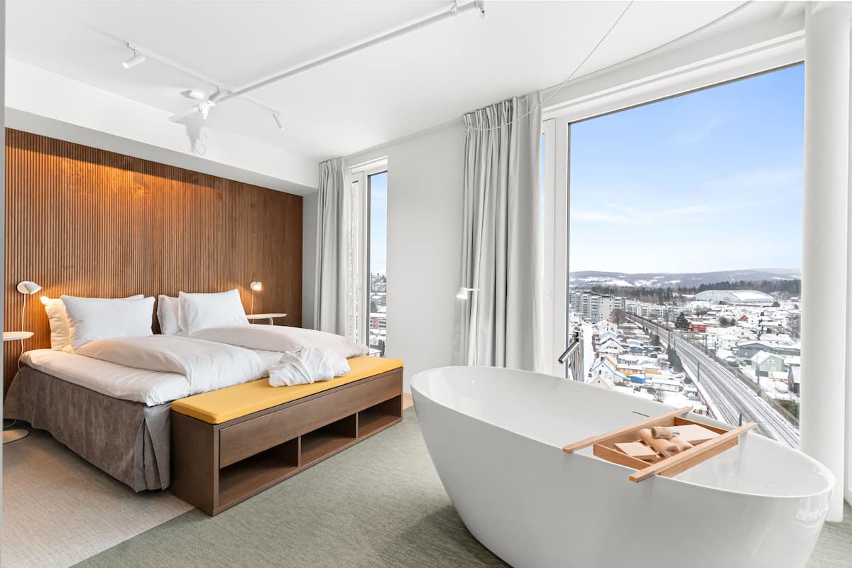 Un dormitorio moderno con una cama grande, un cabecero de madera, un ventanal con vistas a la ciudad y una bañera independiente.