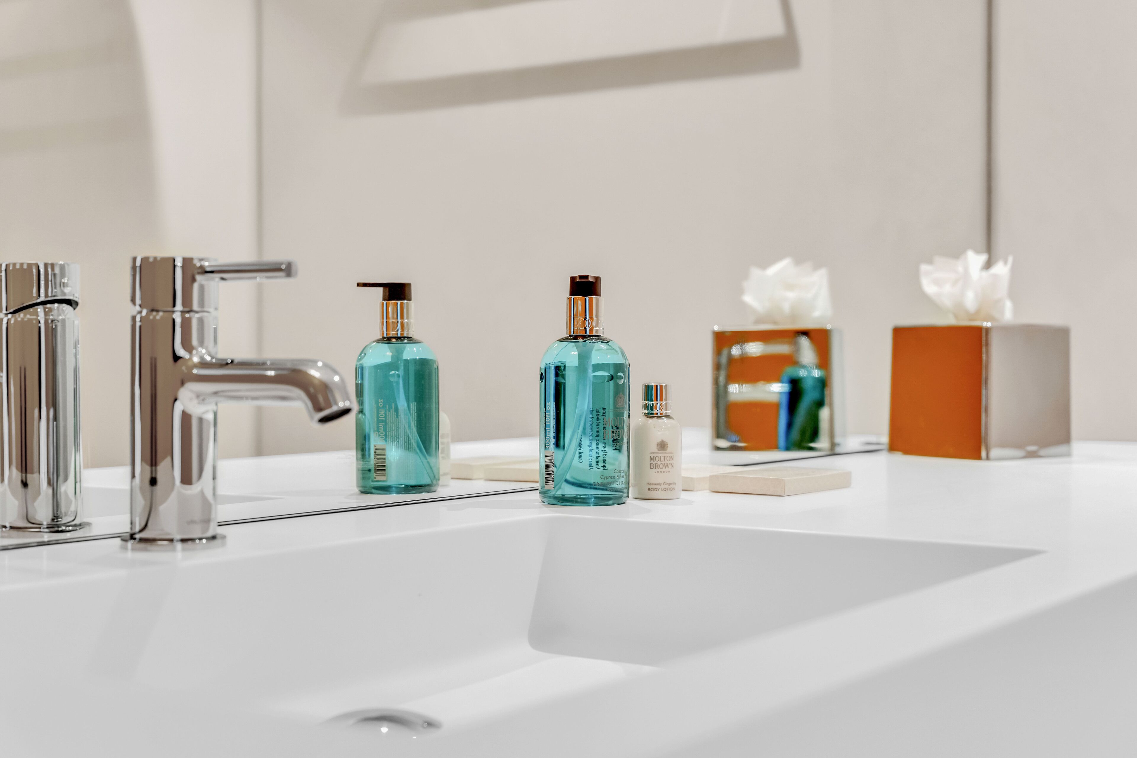 Un baño con un lavamanos blanco, dos grifos cromados y tres botellas de jabón líquido.