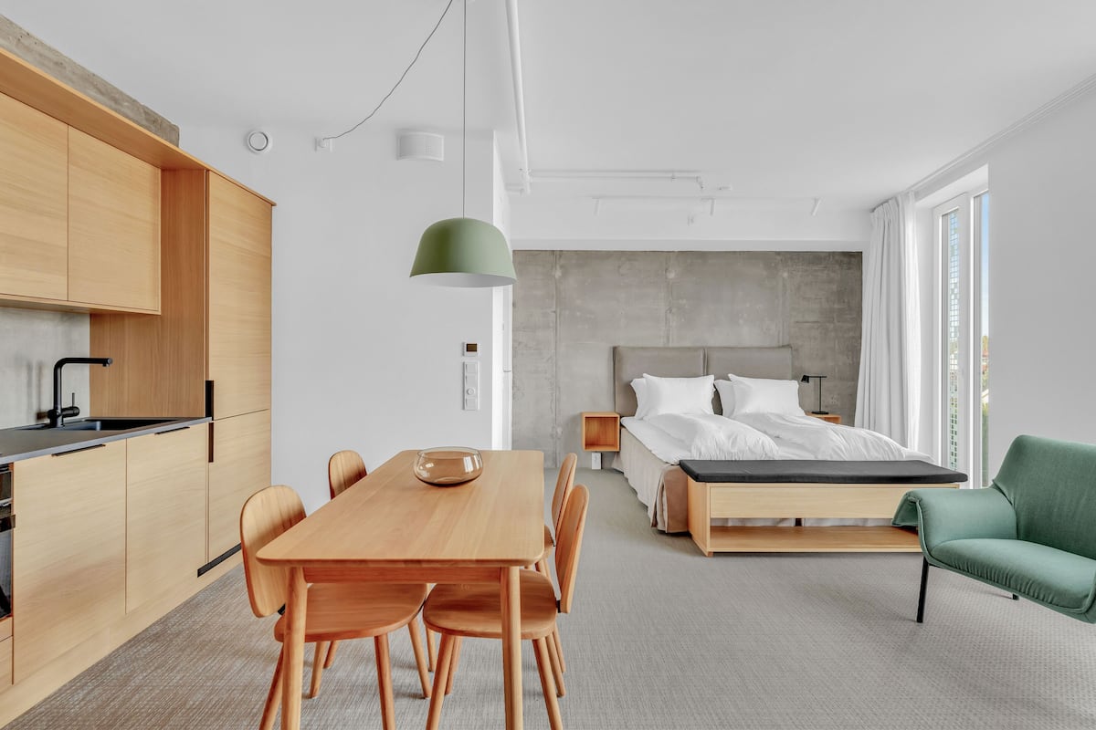Una habitación de hotel moderna con una mesa de comedor de madera, una silla verde, una cama con ropa blanca y un ventanal grande.