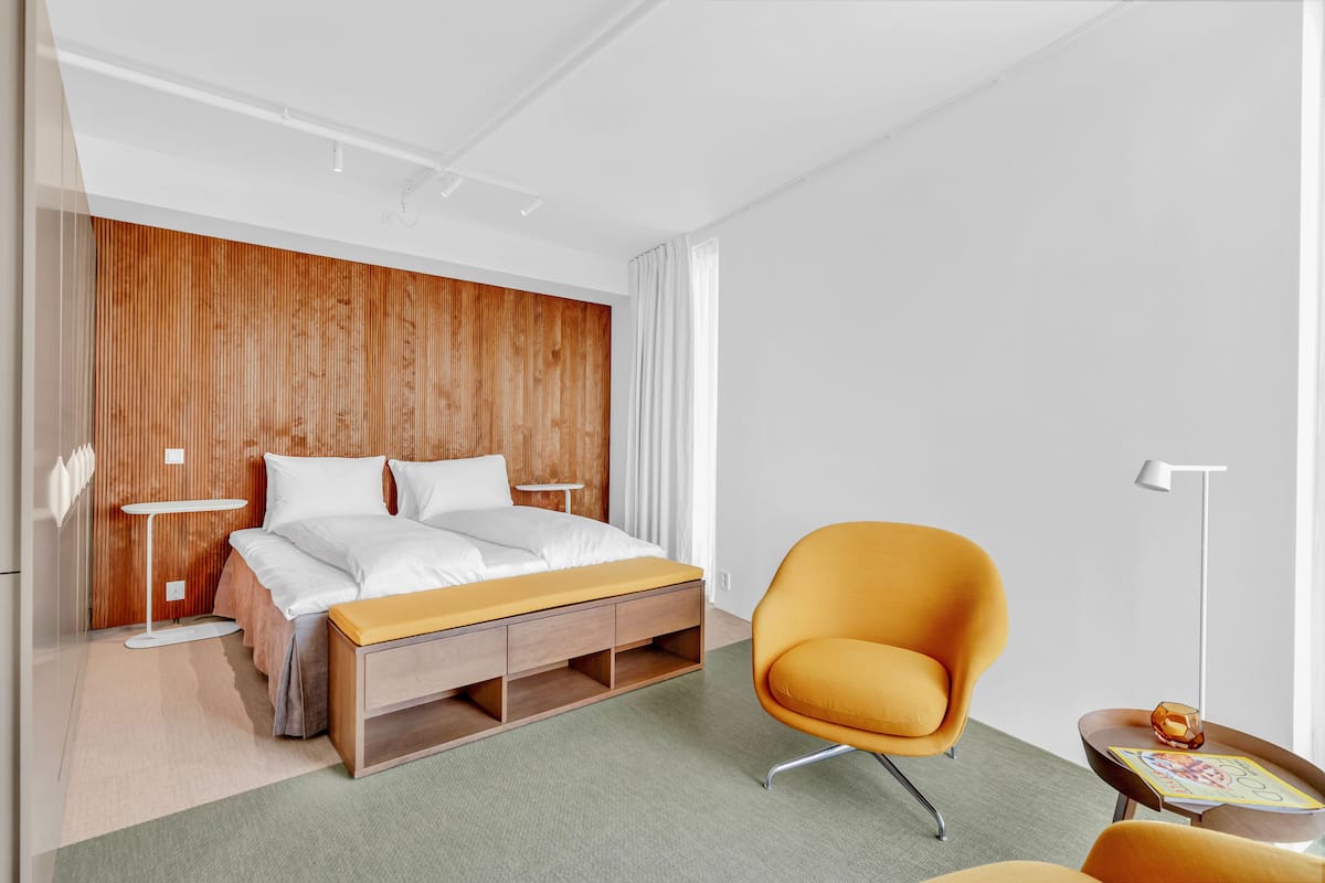 Una habitación de hotel moderna con un cabecero de madera, una cama con ropa blanca, un sillón color mostaza y una mesita de noche con una revista.