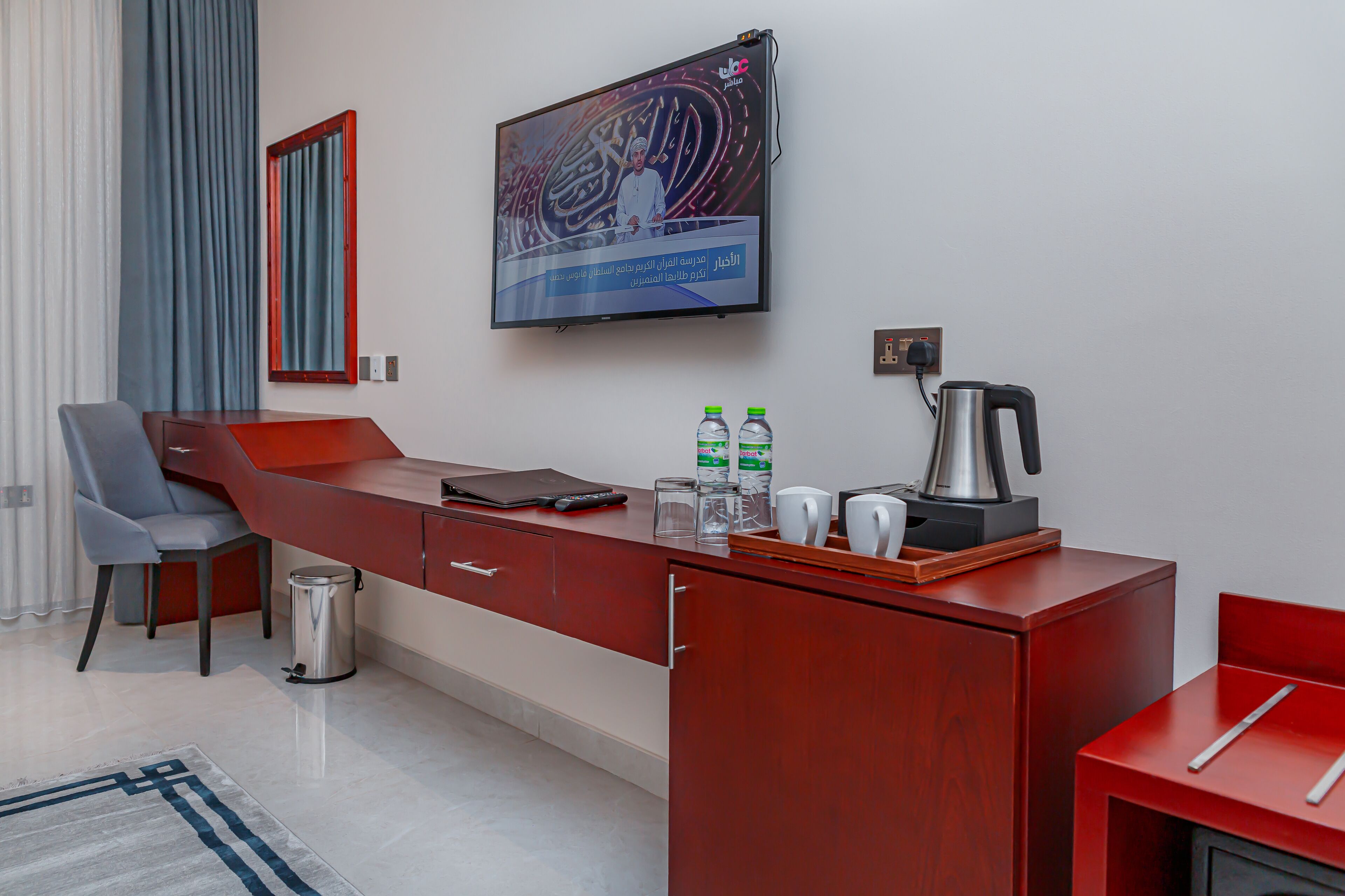 A modern hotel room with a long red desk, a television mounted on the wall, a coffee maker, and a trash can.